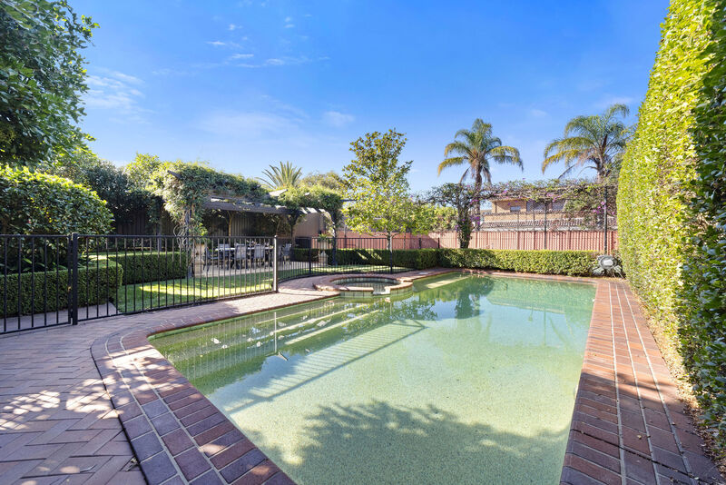 In-ground swimming pool with sandstone coping and a timber pergola, framed by established hedging